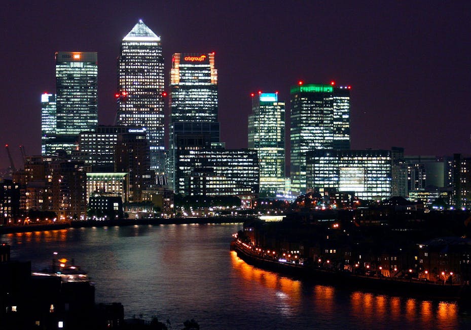 Nighttime city skyline featuring a cluster of illuminated modern skyscrapers with glass facades reflecting lights, including the tall, triangular-topped building in the center, with red lights on top