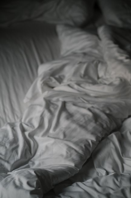 The image shows a neatly made bed with a wooden headboard in a modern indoor setting, featuring a plain white fitted sheet and two large white pillows arranged upright at the head of the bed. The pill